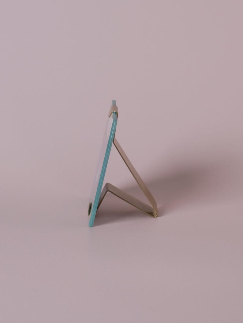 Op de afbeelding: Een kleine, rechthoekige fotolijst met een heldere glazen ruit en een lichtblauwe rand. Het frame wordt ondersteund door een zilverkleurige metalen standaard, ideaal voor het weergeven van foto's of kunstwerken. Het frame is ongeveer 10 cm hoog.