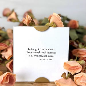 Op de afbeelding: Een witte kaart met een gouden accent, met de quote "be happy in the moment, that's enough. Each moment is all we need, not more." van Moeder Teresa. De kaart is omgeven door perzikroze gedroogde rozen.