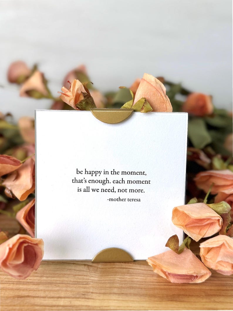 Op de afbeelding: Een witte kaart met een gouden accent, met de quote "be happy in the moment, that's enough. Each moment is all we need, not more." van Moeder Teresa. De kaart is omgeven door perzikroze gedroogde rozen.
