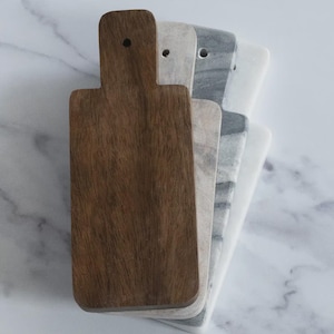 May include: A set of four cutting boards, one brown wood and three marble, stacked on top of each other. The boards have a handle on the top and are all the same size.