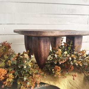 May include: A dark brown wooden riser with a flat square top and four thick, tapered legs. The wood features a visible grain pattern and a smooth, polished finish. The riser is elevated, providing a stable platform for display purposes.