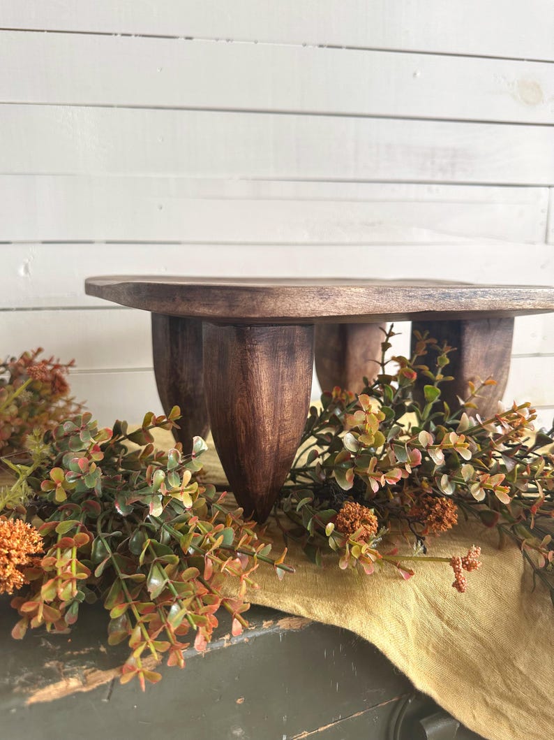 May include: A dark brown wooden riser with a flat square top and four thick, tapered legs. The wood features a visible grain pattern and a smooth, polished finish. The riser is elevated, providing a stable platform for display purposes.