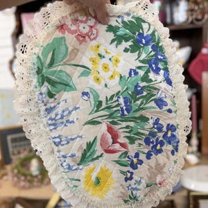 Op de afbeelding: Een ovale, doorgestikte stoffen mat met een bloemenpatroon van blauwe, gele, roze en groene bloemen op een beige achtergrond. De mat is afgewerkt met een brede, crèmekleurige kanten rand die langs de gehele buitenkant van het stuk loopt.