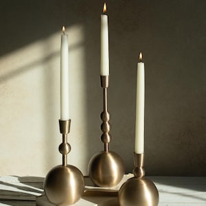 May include: Three gold candle holders with white candles. The candle holders are round and have a textured design. The candles are lit and the flames are visible.