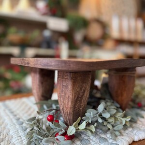 May include: A dark brown wooden riser with a flat square top and four thick, textured legs. The riser is positioned over a decorative arrangement of green foliage and small red berries, showcasing a natural wood grain finish throughout the piece.