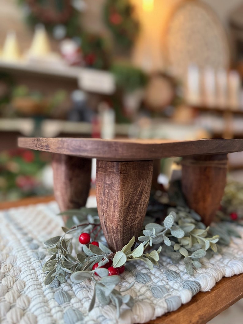 May include: A dark brown wooden riser with a flat square top and four thick, textured legs. The riser is positioned over a decorative arrangement of green foliage and small red berries, showcasing a natural wood grain finish throughout the piece.