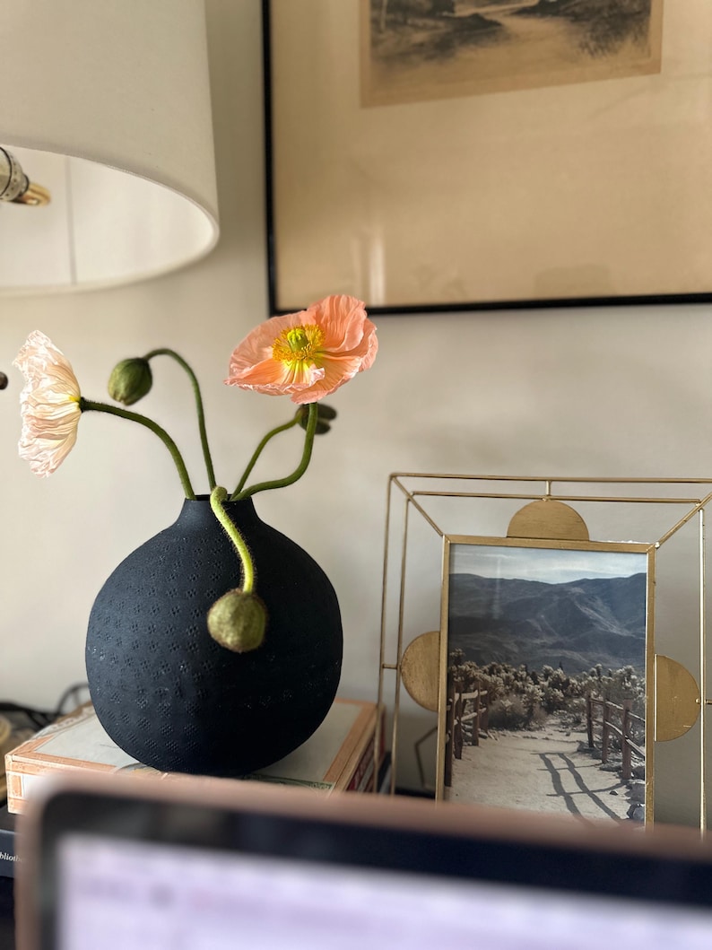 May include: A black, textured vase holding peach-colored poppies with green stems and buds. A gold-colored framed picture of a mountain landscape is to the right of the vase. The vase is sitting on top of a stack of books.