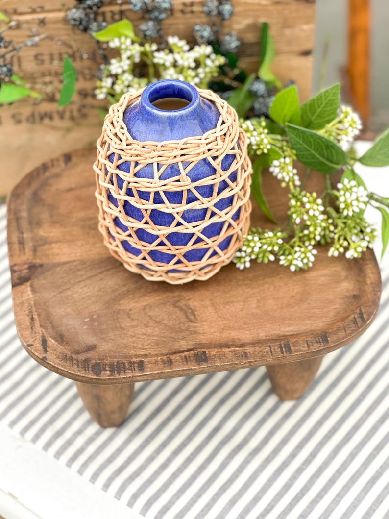 May include: A small, cobalt blue ceramic vase with a narrow neck, encased in a decorative, light-colored woven rattan lattice overlay. The vase features a smooth, glossy glaze finish and sits upright, showcasing a rounded, bulbous body shape.