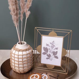 May include: A decorative arrangement featuring a gold-toned metal picture frame with a botanical print, a white vase with a woven design, and a set of wooden peace sign ornaments. The items are displayed on a bronze-colored tray.