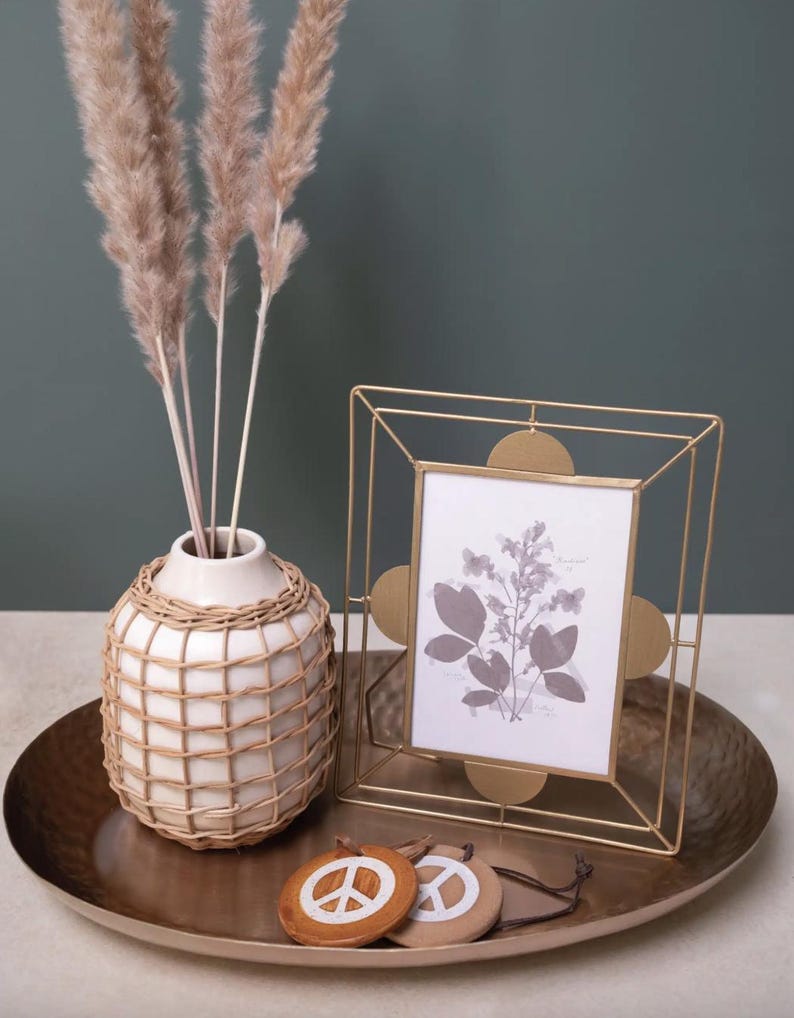 May include: A decorative arrangement featuring a gold-toned metal picture frame with a botanical print, a white vase with a woven design, and a set of wooden peace sign ornaments. The items are displayed on a bronze-colored tray.