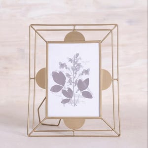 May include: A gold-colored metal picture frame with a geometric design. The frame has a rectangular opening with a botanical print inside. The frame has a stand on the back for display.