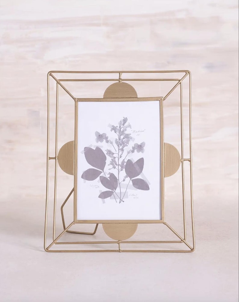 May include: A gold-colored metal picture frame with a geometric design. The frame has a rectangular opening with a botanical print inside. The frame has a stand on the back for display.
