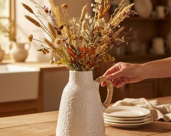 May include: A white ceramic pitcher with a textured, speckled surface and a wrapped rattan handle. The pitcher is filled with a dried floral arrangement featuring various grasses, wheat stalks, and small brown and yellow wildflowers.