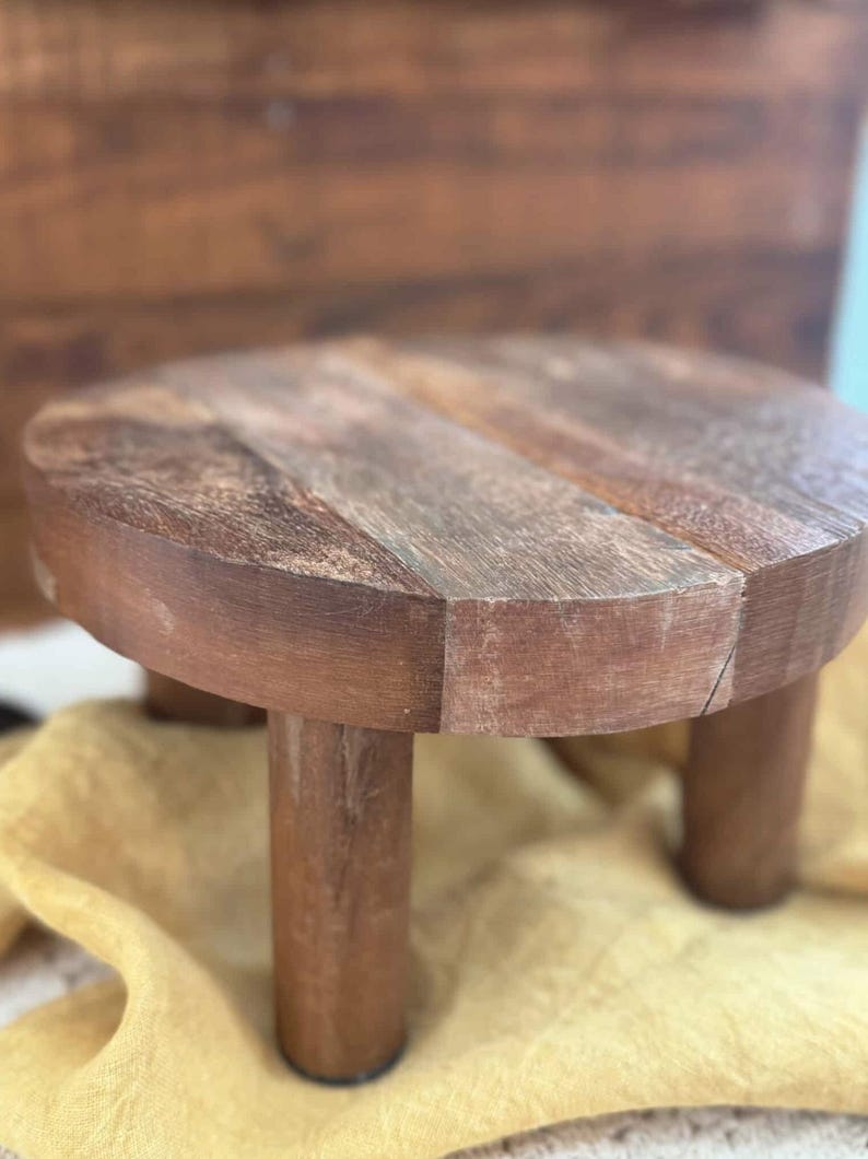 Puede incluir: Un peque&ntilde;o soporte redondo de madera para plantas con tres patas cil&iacute;ndricas. El soporte est&aacute; hecho de madera marr&oacute;n oscuro con vetas visibles y un acabado natural. El soporte est&aacute; dise&ntilde;ado para elevar plantas u otros objetos decorativos.