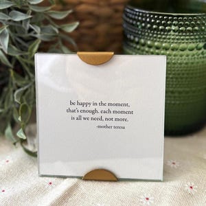 Op de afbeelding: Een vierkante glazen fotolijst met een witte inzet met de quote "be happy in the moment, that's enough. each moment is all we need, not more." van Moeder Teresa. De lijst heeft goudkleurige metalen accenten.