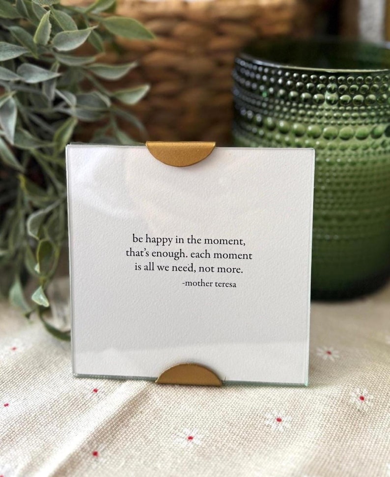 Op de afbeelding: Een vierkante glazen fotolijst met een witte inzet met de quote "be happy in the moment, that's enough. each moment is all we need, not more." van Moeder Teresa. De lijst heeft goudkleurige metalen accenten.