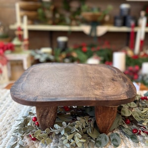 May include: A square wooden riser with a dark brown finish and a smooth, flat top surface. The riser is supported by four short, sturdy, carved wooden legs. The wood grain is visible across the top and the legs of this functional display stand.