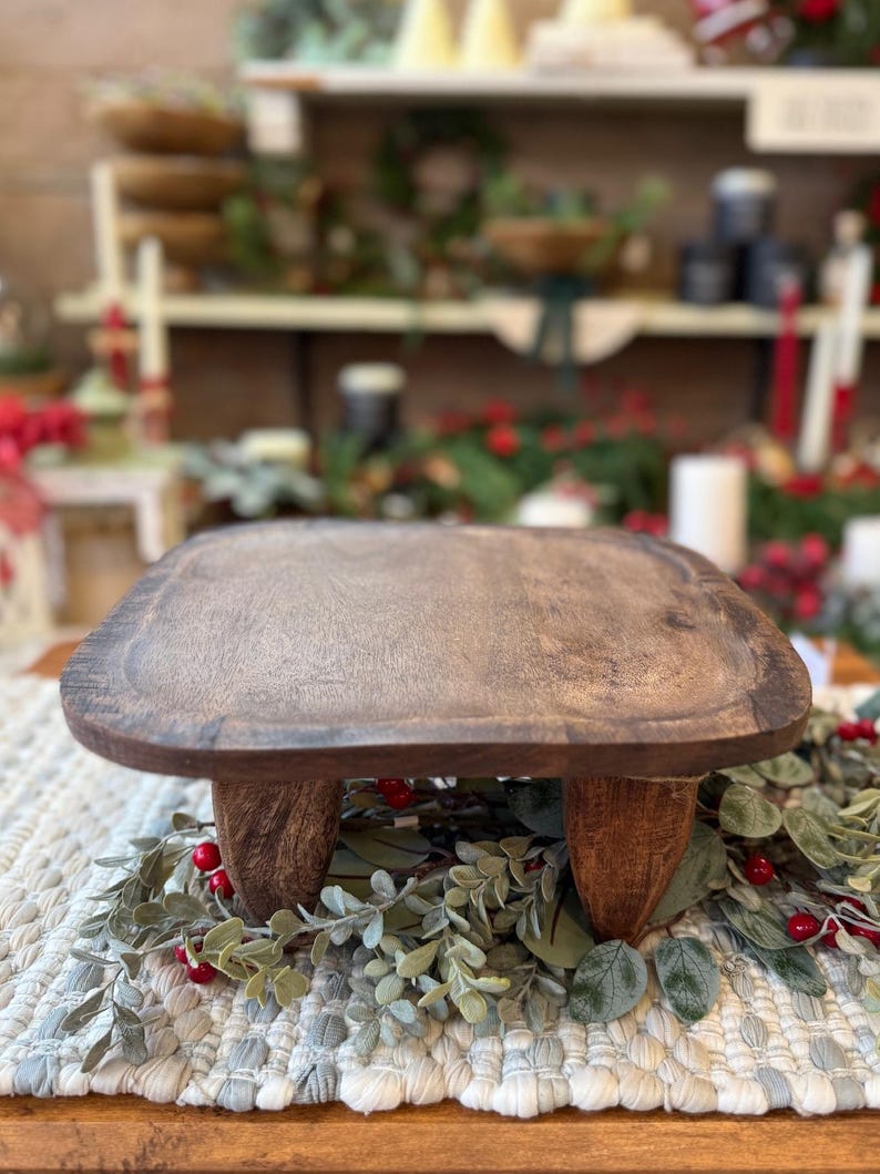 May include: A square wooden riser with a dark brown finish and a smooth, flat top surface. The riser is supported by four short, sturdy, carved wooden legs. The wood grain is visible across the top and the legs of this functional display stand.