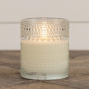 May include: A lit, ivory-colored candle in a clear glass jar. The jar has a textured design of small, raised dots around the top and bottom. The candle sits on a wooden surface, and the background is a white wall.