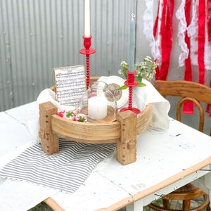 May include: A rustic wooden tray with decorative wooden legs holds a candle, flowers, and a small glass cloche. Two red candle holders with white and blue candles sit on the tray. A striped runner and sheet music are also on the table.