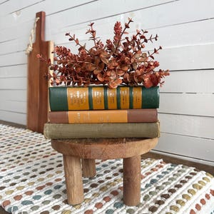 Puede incluir: Un peque&ntilde;o taburete de madera sostiene una pila de libros antiguos con cubiertas verdes, marrones y amarillas, coronada con eucalipto marr&oacute;n rojizo. Una tabla de madera para cortar cuelga en el fondo. Un camino de mesa estampado est&aacute; en primer plano.