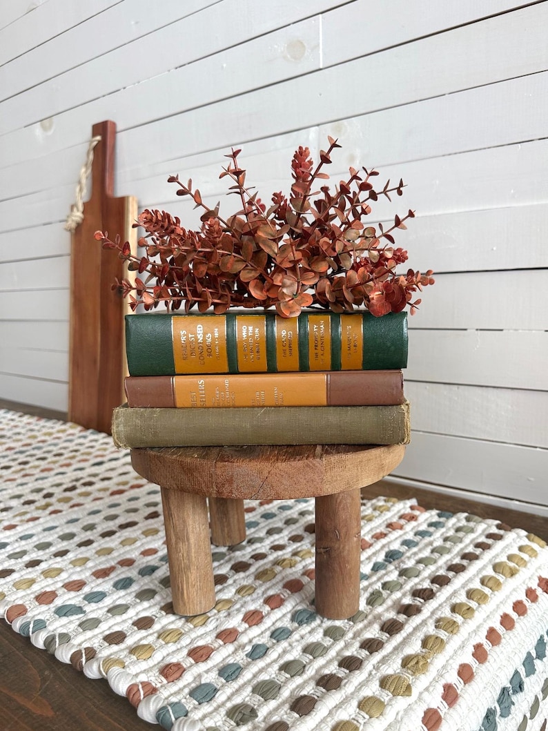 Puede incluir: Un peque&ntilde;o taburete de madera sostiene una pila de libros antiguos con cubiertas verdes, marrones y amarillas, coronada con eucalipto marr&oacute;n rojizo. Una tabla de madera para cortar cuelga en el fondo. Un camino de mesa estampado est&aacute; en primer plano.