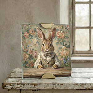 Op de afbeelding: Een ingelijste print met een keurige konijn in een vest en vlinderdas, tegen een bloemenachtergrond. Het kunstwerk wordt weergegeven in een heldere glazen lijst met messingkleurige houders, rustend op een verweerd wit houten oppervlak.