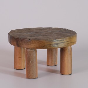 Puede incluir: Un peque&ntilde;o taburete redondo de madera con una parte superior texturizada y desgastada y cuatro patas cil&iacute;ndricas. El taburete est&aacute; hecho de madera clara con un acabado natural y tiene aproximadamente 25 cm de di&aacute;metro.