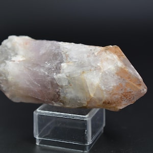Pode incluir: Um cristal de quartzo transparente com uma tonalidade rosa e laranja. O cristal está em um pequeno suporte transparente.