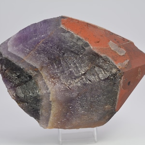 Könnte beinhalten: Ein großer Amethystkristall mit einer natürlichen, rauen Oberfläche. Der Kristall ist tiefviolett mit einer rotbraunen Beschichtung auf einer Seite. Der Kristall befindet sich auf einem weißen Hintergrund.