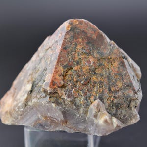 Könnte beinhalten: Ein natürlicher Quarz-Kristall mit pyramidenförmiger Gestalt. Der Kristall weist eine Mischung aus Farben auf, darunter rotbraune, graue und gelbgrüne Farbtöne. Die Oberfläche hat ein raues, strukturiertes Aussehen, das seinen rohen, unpolierten Zustand zeigt. Der Kristall ist ein einzigartiges Exemplar.