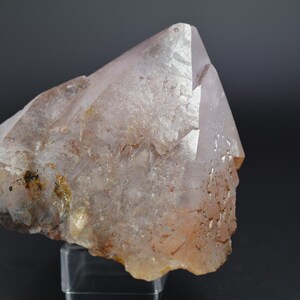 Pode incluir: Um grande cristal de quartzo transparente com uma tonalidade rosa e uma superfície áspera e natural. O cristal tem uma forma pontiaguda e triangular e está parcialmente coberto por inclusões marrons.