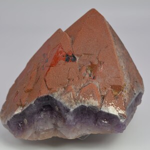 Könnte beinhalten: Eine große, natürliche Kristallformation mit einem rotbraunen Oberteil und einer Basis aus lila Amethyst. Der Kristall hat eine raue, strukturierte Oberfläche und eine einzigartige, eckige Form, ideal zur Präsentation.