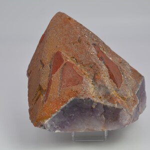 Könnte beinhalten: Ein großer, unregelmäßig geformter Amethystkristall mit einer violetten Farbe. Der Kristall hat eine raue, strukturierte Oberfläche und ist teilweise mit einer Schicht aus rotbraunem Material bedeckt.