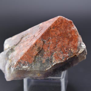 Könnte beinhalten: Natürliches Mineralpräparat mit einer rotbraunen, eckigen Kristallformation. Der Kristall hat eine raue, strukturierte Oberfläche mit grauen und weißen Bereichen. Das Mineral wird auf einem transparenten Ständer vor dunklem Hintergrund ausgestellt.