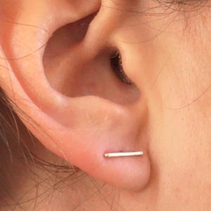 May include: A close-up of a person's ear with a small, silver bar earring. The earring is simple and minimalist, and it is worn in the lobe of the ear.