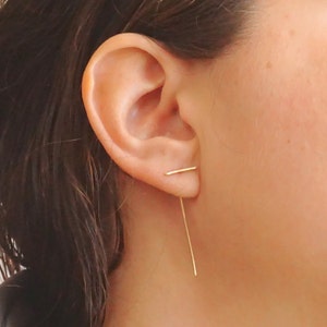 May include: A close-up of an ear with gold bar earrings. One bar is horizontal, on the earlobe, and the other hangs vertically. The earrings have a minimalist design. The earrings are made of gold.