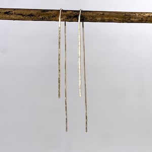 Op de afbeelding: Drie lange, dunne, gouden oorringen die aan een houten stok hangen. De oorringen zijn allemaal recht en hebben een gehamerde textuur.