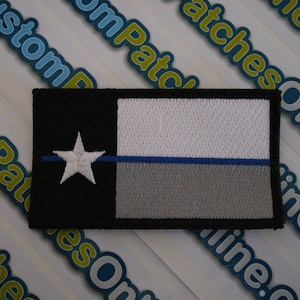 Op de afbeelding: Een geborduurde patch in zwart, wit en grijs met een blauwe lijn en een witte ster. De patch toont de vlag van Texas met een dunne blauwe lijn in het midden.