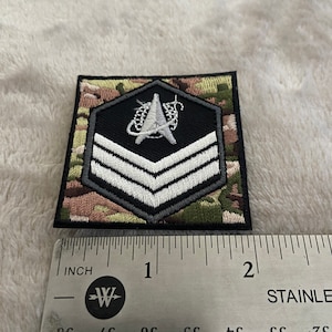 May include: A black and white embroidered patch with a white chevron design and a space shuttle logo. The patch is on a camouflage fabric background. The patch is approximately 1 inch wide.