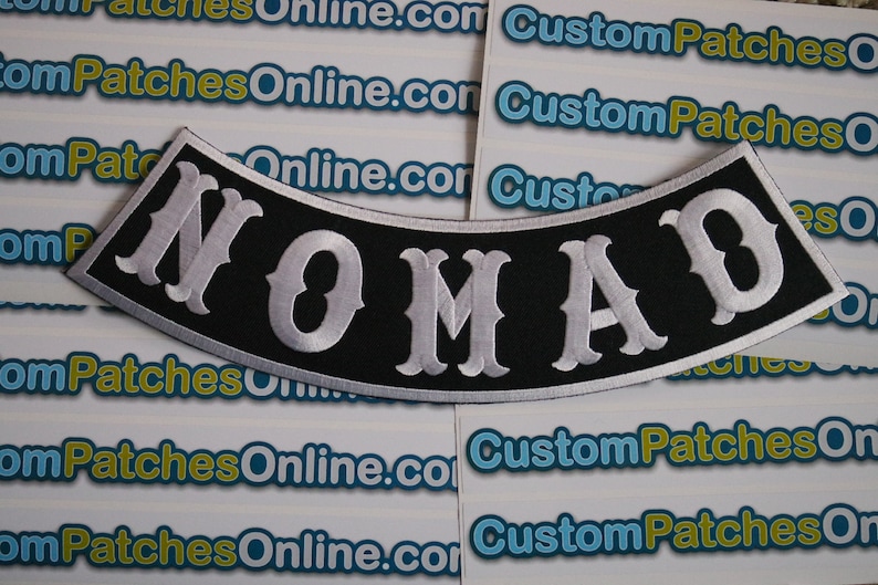 May include: A black and white embroidered patch with the word "NOMAD" in white lettering on a black background. The patch is curved and has a white border.