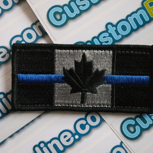 May include: Black and grey embroidered patch with a thin blue line across the middle. The patch features a black maple leaf on a grey background. The patch is surrounded by a black border.