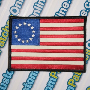 May include: A close-up of a small, embroidered patch featuring the Betsy Ross flag, the first American flag with 13 stars in a circle, representing the original 13 colonies. The flag is red, white, and blue with a black border.