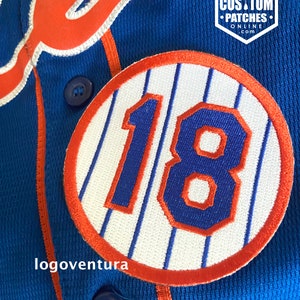 Może przedstawiać: Niebieska koszulka baseballowa z pomarańczowo-białym logo "Mets" z przodu. Koszulka ma biało-pomarańczową naszywkę w paski z numerem "18" w kolorze niebieskim na klatce piersiowej. Koszulka zapinana jest na guziki i ma metkę logoventura.