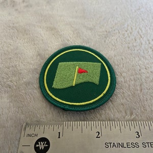 May include: A green and yellow embroidered patch with a golf green and a red flag in the center. The patch is approximately 1.5 inches in diameter.