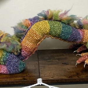 May include: A handmade, colorful dragon plushie with a rainbow color scheme. The dragon features a detailed crochet body, fluffy accents, and a flower-shaped eye. The dragon is resting on a wooden surface.