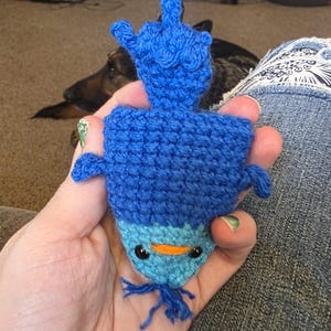 May include: A handmade crochet blue creature with a lighter blue belly, orange beak, and black eyes. The creature has a long, upright appendage and small side fins. The item is held in a person's hand.