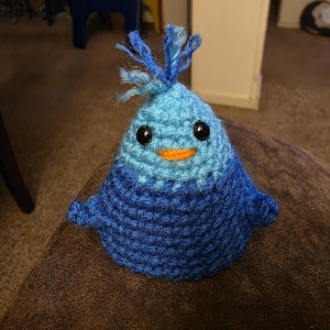 May include: A handmade crochet creature in shades of blue, with black button eyes and an orange mouth. The plush toy has a tuft of blue yarn on top and small arms. This whimsical item is perfect for a child's room or as a unique gift.
