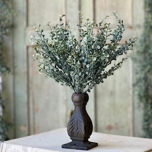 May include: A bouquet of faux eucalyptus sprigs in a dark brown, ornate vase. The vase is on a white book with text.