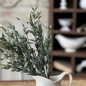 May include: A white ceramic pitcher with a wide mouth and a handle, filled with a sprig of green leafy branches.